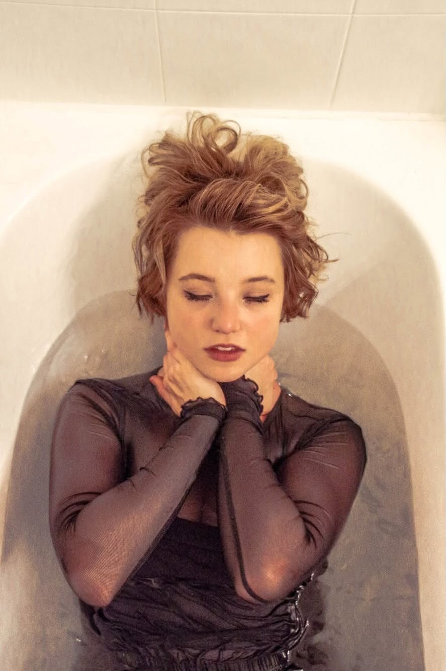 Forest Evergreen van Eeden lying in a bath of water with eyes closed, wearing a sheer plum top